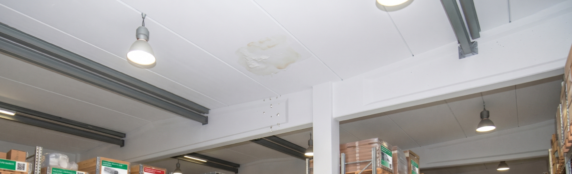 Warehouse ceiling with visible water stains and moisture damage, signaling flat roof leaks above a storage area that require inspection.